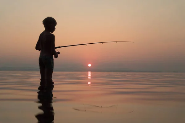 Jongen vissen met een hengel staande in het water in de avond bij zonsondergang — Stockfoto