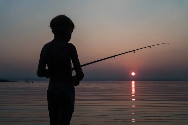 Niño pescando con una caña de pescar de pie en el agua por la noche al atardecer — Foto de Stock