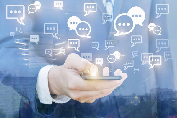 A businessman is holding a mobile phone from which different symbols are illuminated. The concept is the usage of social networks