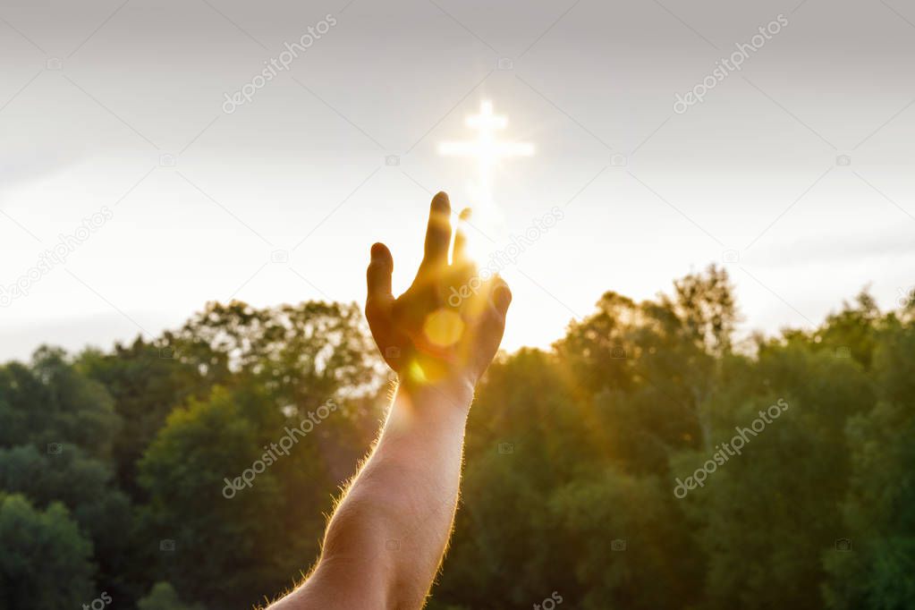 A hand is reaching a glowing cross image which is situated in the sky above the green summer trees. The concept is the strength of feith.