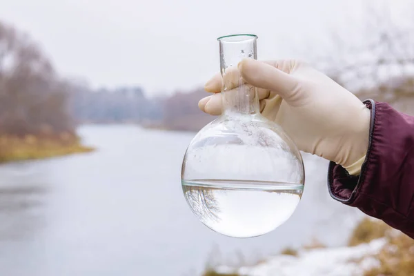 A retort with river water sample inside in the laboratorian\'s hand at the countryside landscape background. The concept is the environment protection