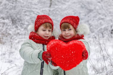 İki küçük gülümseyen kız ikiz benzer parlak giysili kar taneleri uçan tarafından çevrili büyük konforlu kırmızı kalp kış arka plan, ellerinde gösterilen.