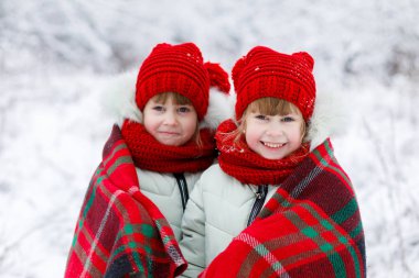 Küçük kız kardeş ikizler gülümseyen giyinmiş olarak parlak kıyafetleri birlikte ayakta karlı arka plan bulanık, sıcak renkli ekose kaplı