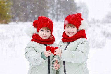 İki mutlu sıcak gülümseyen kız ikiz giyinmiş renkli elbise karlı kış orman arka plan, ellerinde iki küçük parlak örme kalp gösterilen. Sevgililer günü kutlama bir kavramdır.
