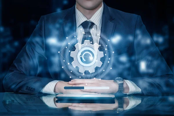 A businessman is sitting at the table with the digital update sign system of networking connections and cogwheel above the device at the blurred background. The concept of networking system update.