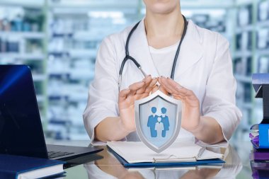 Genç doktor bir kalkan kişi veya aile üyeleri simgesi içinde Grup ile yukarıda koruyucu jest olarak ellerini tutarak bir masada oturuyor. Aile Hekimliği ve hayat ve sağlık sigortası kavramı