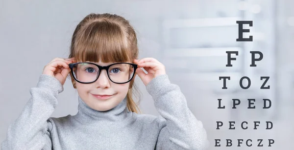 A smiling child in glasses is standing with checking table with letters behind her at the blurred medical room background. The concept of ophthalmologic diagnostic, treatment and correction.