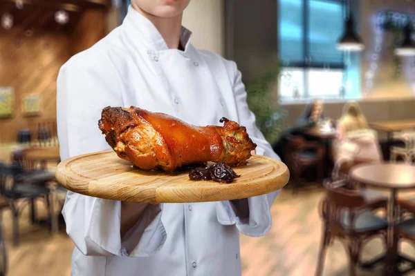 A closeup of a chief cook holding a wooden tray with baked pork and dried fruits on it at the blurred restaurant interior background.