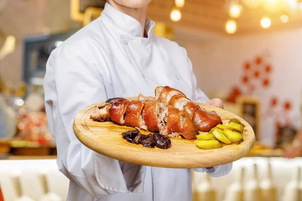 A closeup of chief cook holding tray with combination of prepared meat, potato and dried fruits on it at the blurred restaurant interior background. The concept of eating out or professional cuisine.