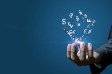 Avuçlarında para birimlerinin sembollerinden oluşan bir dünya tutuyor. Bir el, küresel finans ve ticareti gösteren çeşitli para birimi sembollerini havada tutuyor..