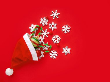 Yeni yıl. Noel Baba şapkası, beyaz kar taneleri ve parlak kırmızı arka planda Noel süsleri. Üst görünüm, alanı kopyala
