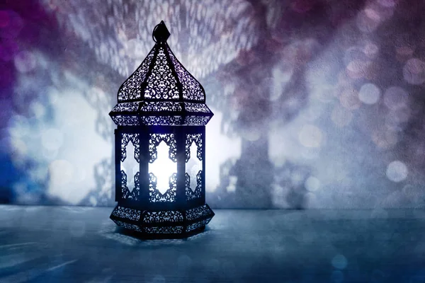 Ornamental Arabic lantern with burning candle glowing at night and glittering blue bokeh lights. Festive greeting card, invitation for Muslim holy month Ramadan Kareem. Blurred party background