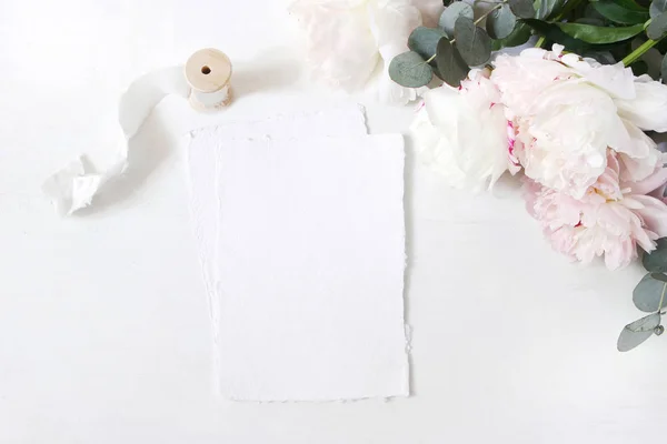 Θηλυκή γάμο ή γενέθλια πίνακα σύνθεση με floral μπουκέτο. Λουλούδια άσπρα και ροζ παιώνιες, ευκάλυπτος και μετάξι κορδέλα. Κενό βαμβάκι χαρτί καρτών mockups, προσκλήσεις. Επίπεδη lay, κορυφαία προβολή. — Φωτογραφία Αρχείου