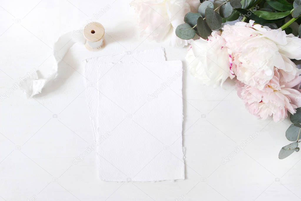 Feminine wedding or birthday table composition with floral bouquet. White and pink peonies flowers, eucalyptus and silk ribbon. Blank cotton paper cards mockups, invitations. Flat lay, top view.