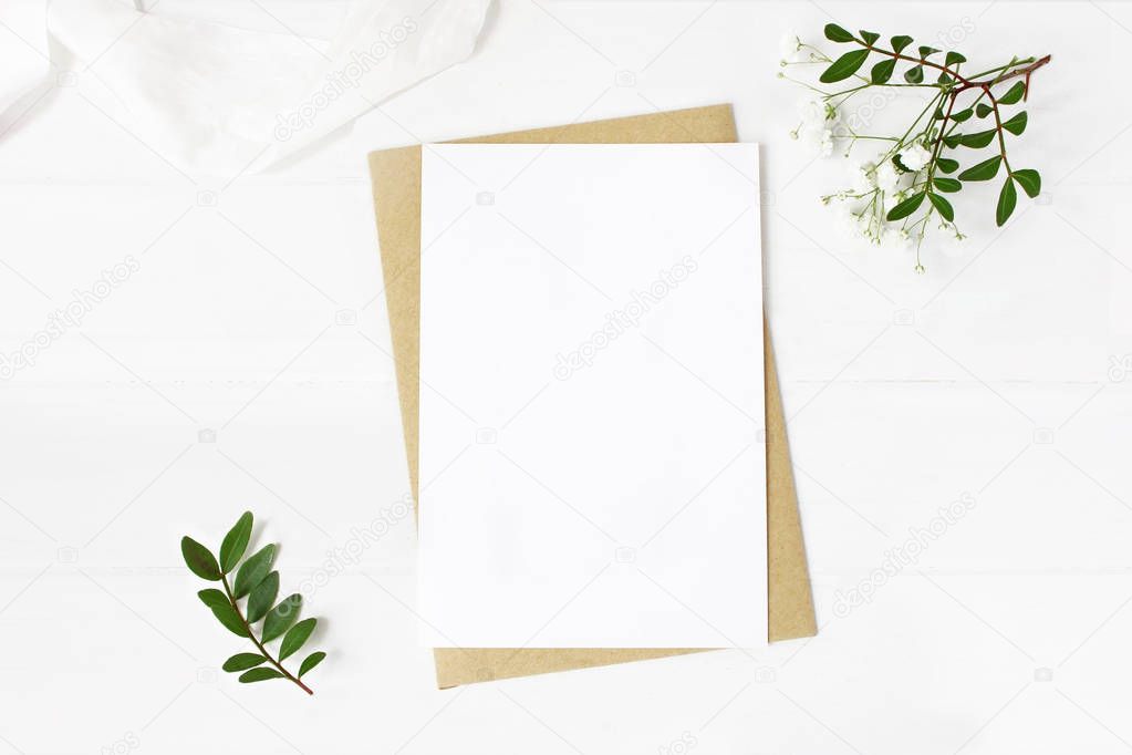 Feminine wedding stationery, desktop mock-up scene. Blank greeting card, craft envelope, babys breath flowers, silk ribbon and lentisk branches, old white wooden table background. Flat lay, top view.