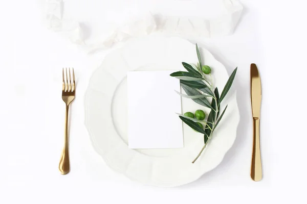 Cenário festivo de verão de mesa com talheres dourados, ramo de oliveira, prato de jantar de porcelana e fita de seda no fundo da mesa branca. Um maquete de cartas em branco. Conceito de menu de casamento ou restaurante mediterrânico — Fotografia de Stock