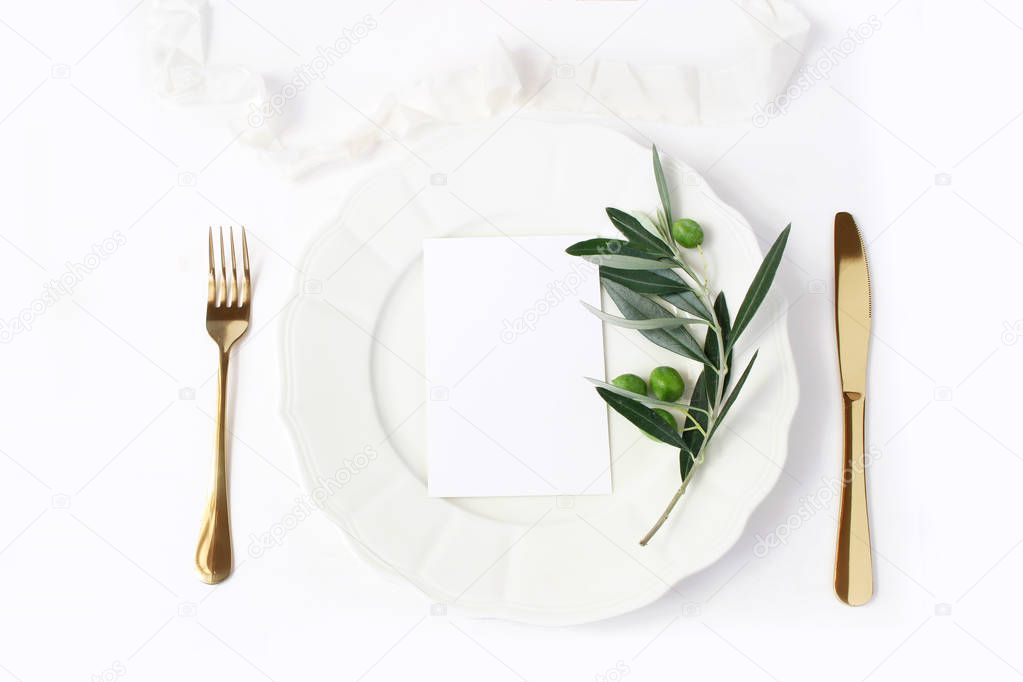 Festive table summer setting with golden cutlery, olive branch, porcelain dinner plate and silk ribbon on white table background. Blank card mockup. Mediterranean wedding or restaurant menu concept