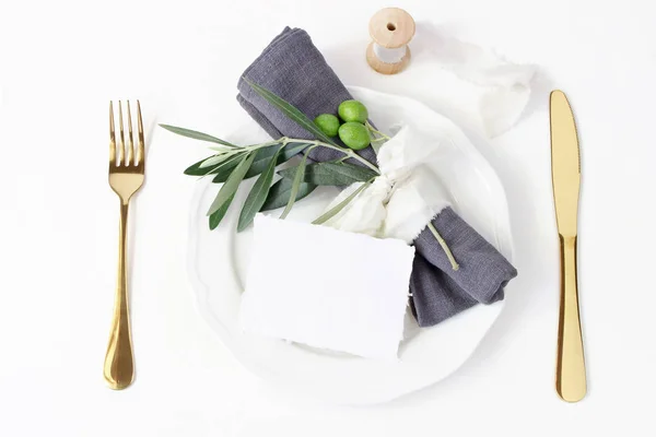 Mesa festiva de verano. Cubertería dorada, rama de olivo, servilleta de lino, plato de porcelana y cinta de seda sobre fondo de mesa blanco. Una maqueta de cartas en blanco. Menú de boda o restaurante mediterráneo — Foto de Stock