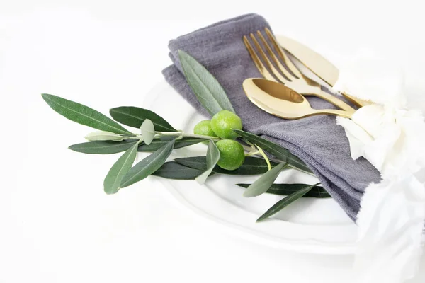 Primer plano de la mesa festiva de verano con cubiertos de oro, rama de olivo, servilleta de lino gris, plato de porcelana y cinta de seda sobre fondo de mesa blanco. Boda mediterránea, restaurante . — Foto de Stock