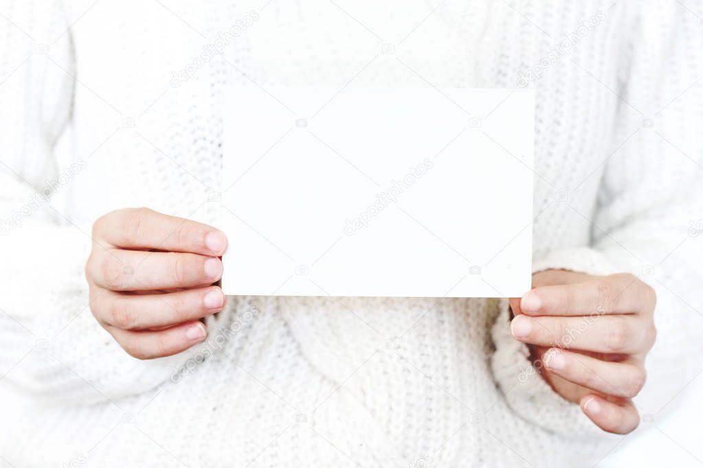 Closeup of womans hands in white knitted sweater holding blank paper card. Greeting card mock-up. Chritsmas winter design. Feminine styled stock photo.