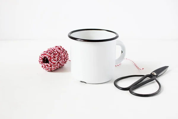 Composition nature morte d'hiver avec tasse à café en métal blanc vierge, ciseaux de vitage et corde cadeau décorative rouge sur fond de table blanche. Photo de stock de style Noël, scène rustique. Modèle de produit . — Photo