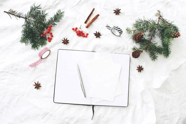 Christmas composition. Blank note pad, greeting cards mock-up scene. Christmas tree branches, red rowan berries, pine cones, washi tapes and cinnamon. White linen tablecloth background. Flat lay, top. — Stock Photo, Image