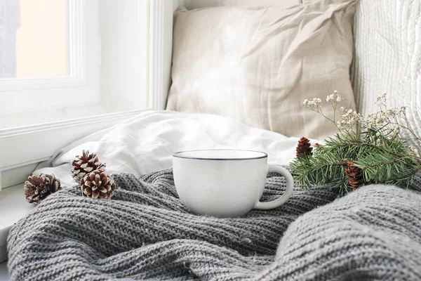 Ζεστό χειμωνιάτικο πρωινό πρωινό στο κρεβάτι ακόμα ζωή σκηνή. Αχνιστό φλιτζάνι ζεστό καφέ, τσάι στέκεται κοντά σε παράθυρο. Χριστουγεννιάτικη ιδέα. Μαξιλάρια, κουκουνάρια και υποκατάστημα δέντρο έλατο σε μάλλινο καρό. — Φωτογραφία Αρχείου