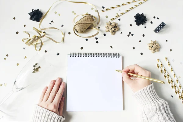 Mains féminines en pull et écriture. Nouvelle résolution d'année, concept de liste de souhaits. Modélisation de lettre en papier vierge. Décoration de fête dorée, confettis et verre à vin sur fond de table blanc. Couché plat, vue du dessus . — Photo