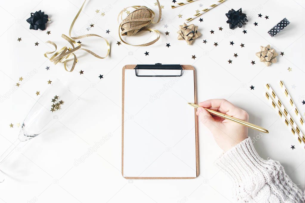 Female hand in sweater and writing. New year resolution, wish list concept. Blank notebook mockup. Golden party decoration, confetti and wine glass on white table background. Flat lay, top view.