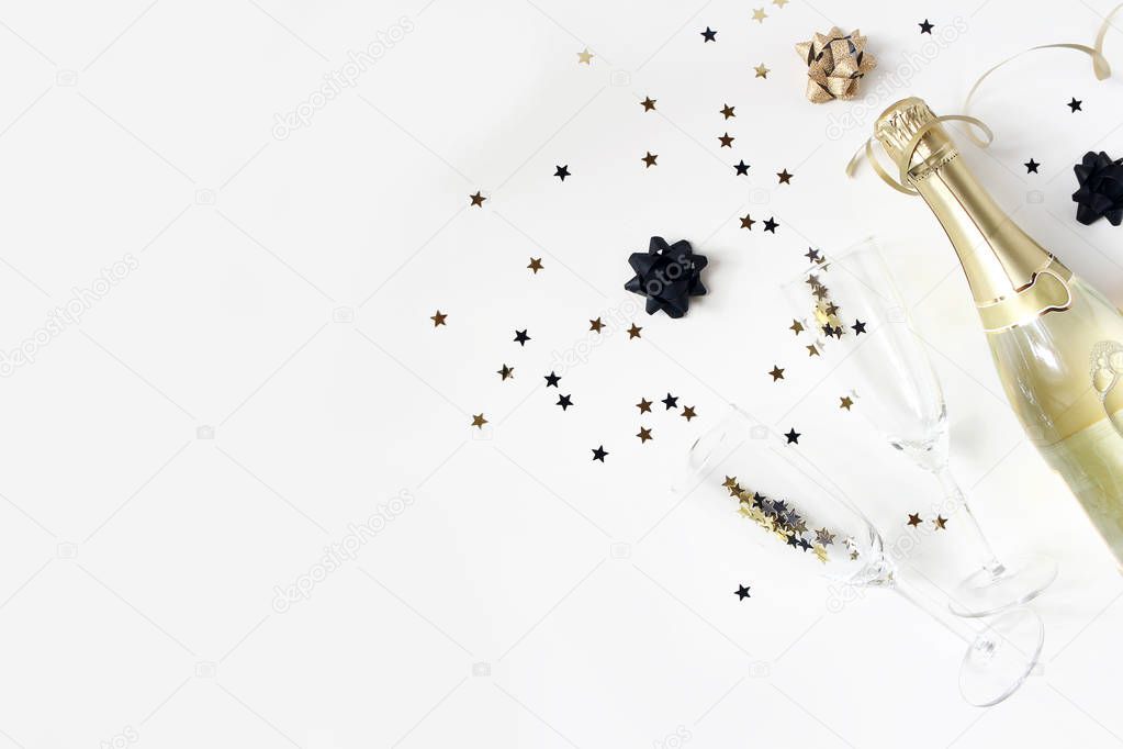 Happy New Year composition. Champagne wine bottle and glasses with golden confetti stars isolated on white table background. Party celebration concept. Flat lay, top view. Empty copy space.