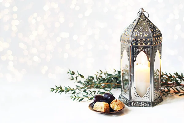 Ramadan Kareem greeting card, invitation. Bronze plate with dates fruit, baklava pastry, burning silver Moroccan, Arabic lantern on white table. Iftar dinner. Glittering lights. Eid ul Adha background — Stock Photo, Image