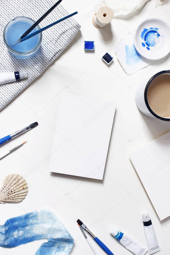 Watercolor, oil, acrylic color paint tubes, brushes, palette, cup of coffee and blank paper card mockups isolated on white table. Art supplies scene. Creative background. Flat lay, top view, vertical.