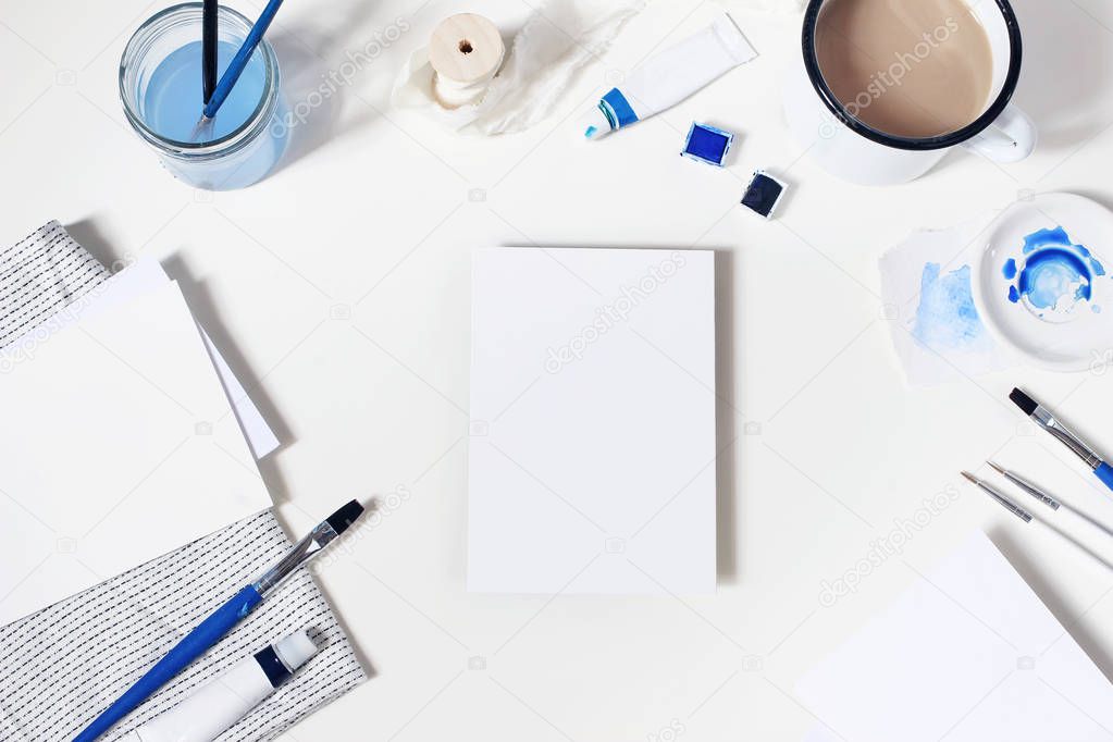 Watercolor, blue oil, acrylic color paint tubes, brushes, palette, cup of coffee and blank paper card mockups isolated on white table. Art supplies. Creative background scene. Flat lay, top view.