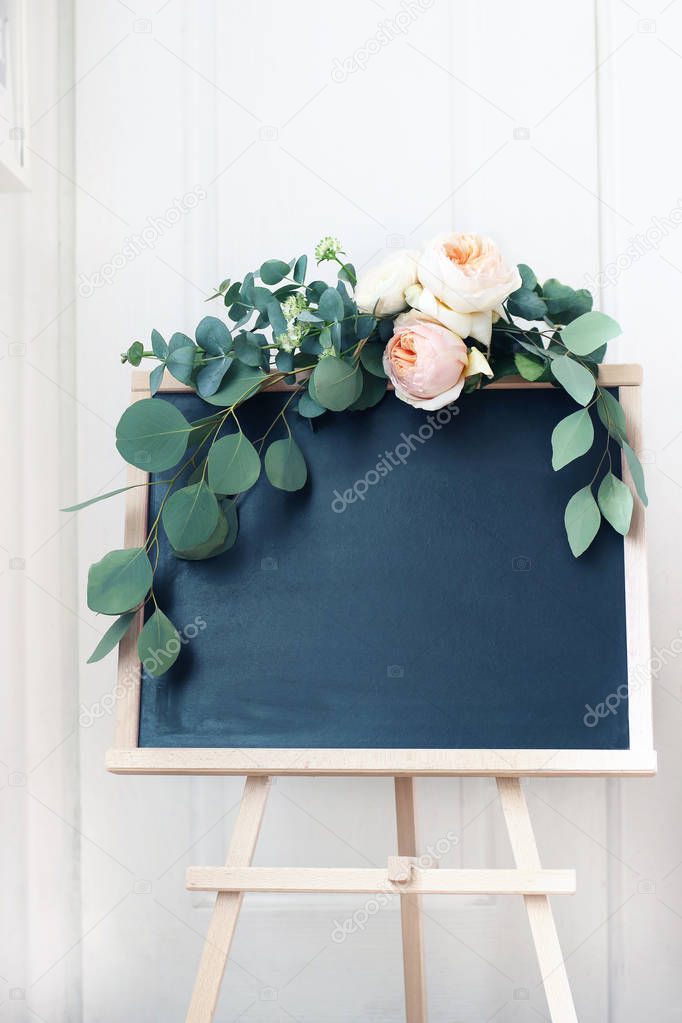 Empty wedding chalkboard sign mockup scene. Floral garland of eucalyptus branches and apricot English roses flowers. Rustic birthday party decoration. Wooden easel with welcome board. Vertical.