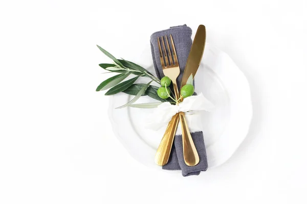 Primer plano de la mesa festiva de verano con cubiertos de oro, rama de olivo, servilleta de lino gris, plato de porcelana y cinta de seda sobre fondo de mesa blanco. Boda mediterránea, restaurante . — Foto de Stock