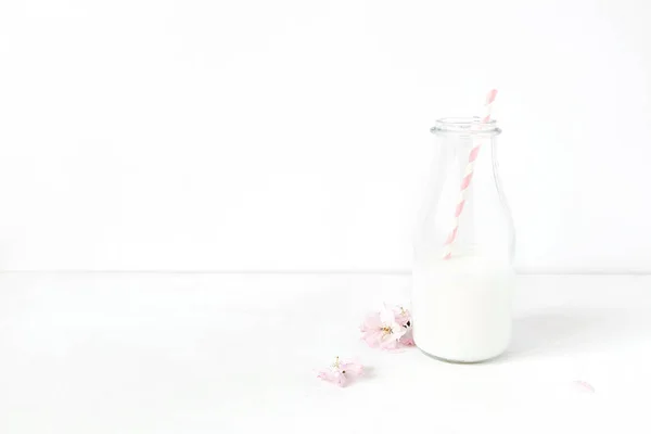 Desayuno fiesta escena de escritorio. Composición con tarro de vidrio de leche, paja de papel para beber rosa y sakura, flores de cerezo japonés sobre fondo de mesa blanco. Primavera, diseño de Pascua . — Foto de Stock