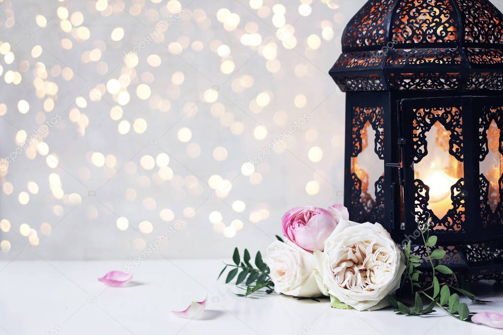 Closeup of vintage black Moroccan, Arabic lantern, glowing candle, green branches, rose flowers and petals on white table background. Greeting card for Muslim holiday Ramadan Kareem, bokeh lights.