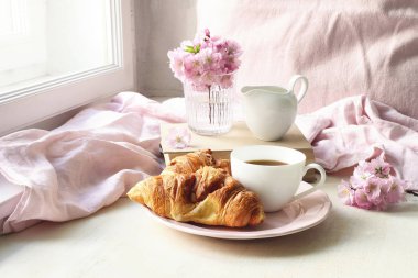 Bahar natürmort sahnesi. Bir fincan kahve, kruvasan böreği, eski kitaplar ve süt sürahisi. Vintage kadınsı tarz fotoğraf. Pembe sakura ile çiçek kompozisyonu, beyaz masada kiraz ağacı çiçekleri.