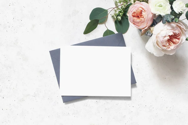 Matrimonio femminile, scena del modello di compleanno. Biglietto d'auguri di carta bianca, busta. Bouquet di foglie di eucalipto, rose rosa arrossite inglesi e fiori di ranuncolo. Sfondo tavolo in cemento. Posa piatta, superiore — Foto Stock