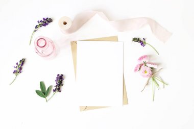 Kadınkırı, masaüstü maket sahnesi. Dikey boş tebrik kartı, zanaat kağıt zarf, ipek kurdele, papatya ve yonca çiçekleri. Beyaz masa arka planı. Düz yatıyordu, üstten bak. Rustik kompozisyon.