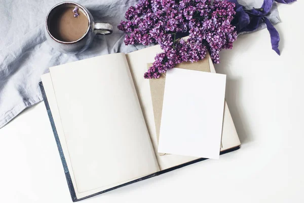 Spring feminine scene, floral composition. Bunch of purple and white lilac flowers, old book, cup of coffee and linen tablecloth on white background. Blank greeting card mockup. Flat lay, top view. — Stock Photo, Image