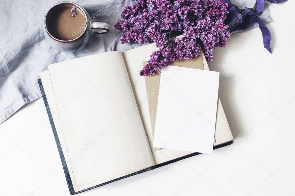 Spring feminine scene, floral composition. Bunch of purple and white lilac flowers, old book, cup of coffee and linen tablecloth on white background. Blank greeting card mockup. Flat lay, top view.