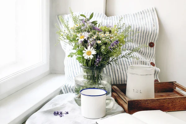夏の静物シーン。エナメルマグ、野生の花の花束とリネンクッション。ヴィンテージフェミニンなスタイルの写真、素朴なデザイン。窓の近くのテーブルの上にコーヒー、ラベンダー、ヒナギクと花の組成物. — ストック写真