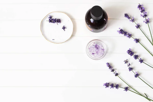 Composizione di bellezza estiva in stile. Essenziale, olio da massaggio, fiori di lavanda viola e sale da bagno su sfondo bianco tavolo di legno. Cosmetici biologici alle erbe, concetto spa. Spazio vuoto, posizione piatta, vista dall'alto — Foto Stock