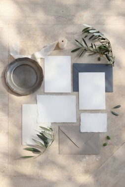 Yaz düğün kırtasiye mock-up sahne. Boş tebrik kartları, zarflar, gümüş tabak, ipek kurdele ve zeytin dalları güneş ışığında. Gölgeleri ile mermer arka plan. Dikey kadınsı düz döşeme, üst görünüm