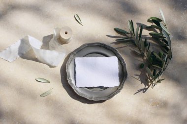 Yaz düğün kırtasiye mock-up sahne. Zeytin dalı, yaprak ve ipek kurdele ile vintage gümüş plaka üzerinde boş pamuk yer kartı. Güneş ışığında mermer arka plan, gölgeler. Düz döşeme, üst görünüm.