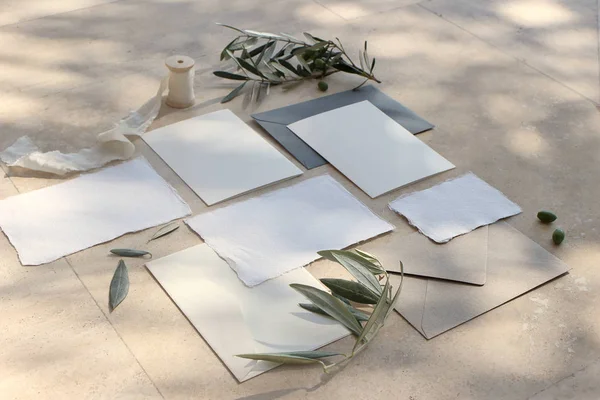 Summer wedding stationery mock-up scene. Blank greeting cards, envelopes, silk ribbon and olive branches and fruit in sunlight. Marble background with shadows. Feminine flat lay, top view.