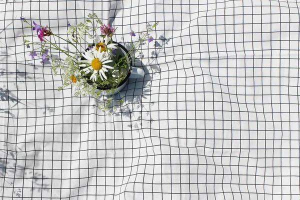 Mug with wild fowers bouquet. Bells, cow parsley and daisies on white checkered blanket, plaid in sunny day. Spring, summer outdoor meal, relaxation concept. Flat lay, top view, empty copy space. — Stock Photo, Image
