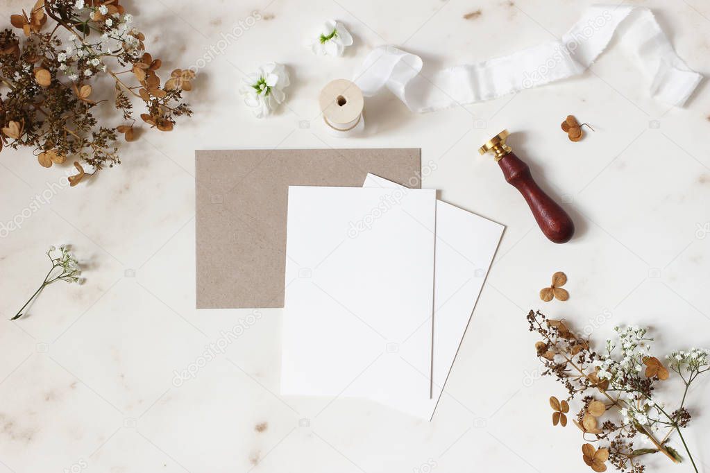 Winter, fall wedding, birthday table composition. Stationery mockup scene. Greeting cards, envelope, dry hydrangea flowers, wax seal stamp and ribbon on marble table backgound. Flat lay, top view.