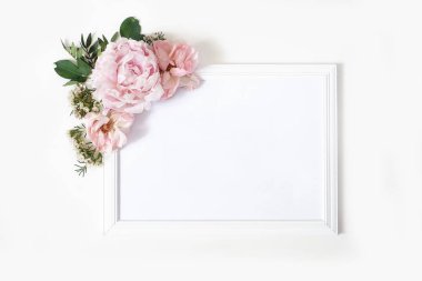 Düğün, doğum günü tabelası maket sahnesi. Boş beyaz ahşap çerçeve. Dekoratif çiçek köşesi. Yeşil yapraklar, pembe şakayık, güller ve balmumu çiçekler. Beyaz masa arka planı. Düz döşeme, üst görünüm.